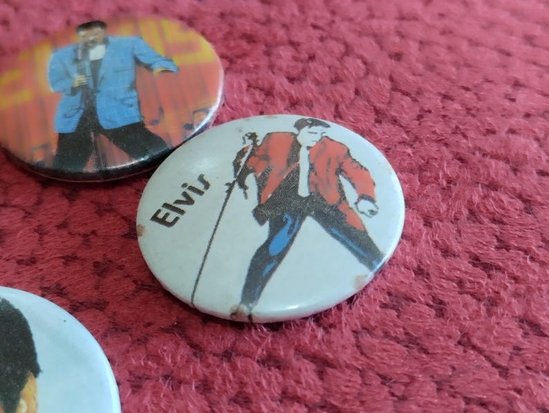 May include: Two round pinback buttons featuring Elvis Presley. The first button has a red, white, and blue background with a black and white image of Elvis Presley in a blue suit. The second button has a white background with a black and white image of Elvis Presley in a red suit. The text "Elvis" is printed in black on the second button.