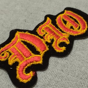 May include: Embroidered patch with the letters "FO" in a stylized, ornate font. The letters are red with yellow outlining, set against a black background. The patch is likely for clothing or accessories.