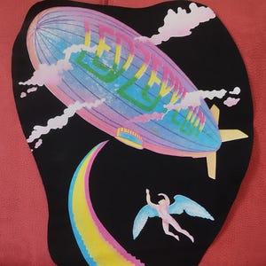 Puede incluir: Un parche de tela negro con una ilustración colorida de un dirigible con la palabra "LED ZEPPELIN" en el lateral. Una escalera de colores arcoíris conduce a una figura alada que vuela en el cielo.