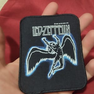Könnte beinhalten: Ein rechteckiger schwarzer Stoffaufnäher mit dem weißen Led Zeppelin-Logo und einer Grafik einer geflügelten Figur in Schwarz-Weiß mit blauen Akzenten. Der Text 1984 Atlantic Recording Corp ist in kleinen weißen Buchstaben oben mittig gedruckt.