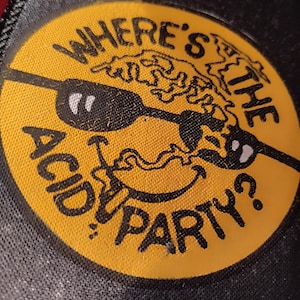 Peut inclure: Un écusson circulaire jaune avec le texte "WHERE'S THE ACID PARTY?" en noir. L'écusson présente une illustration noire d'un visage avec des lunettes de soleil et un motif ondulé. L'écusson est sur un fond noir.