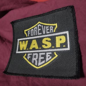 May include: A square black embroidered patch featuring a yellow and white shield design. The text FOREVER is embroidered in white above the yellow text W.A.S.P. inside a white rectangular border, with the word FREE in white below the border.