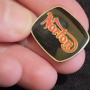 May include: A black and gold square pin with the word "Norton" in red lettering.
