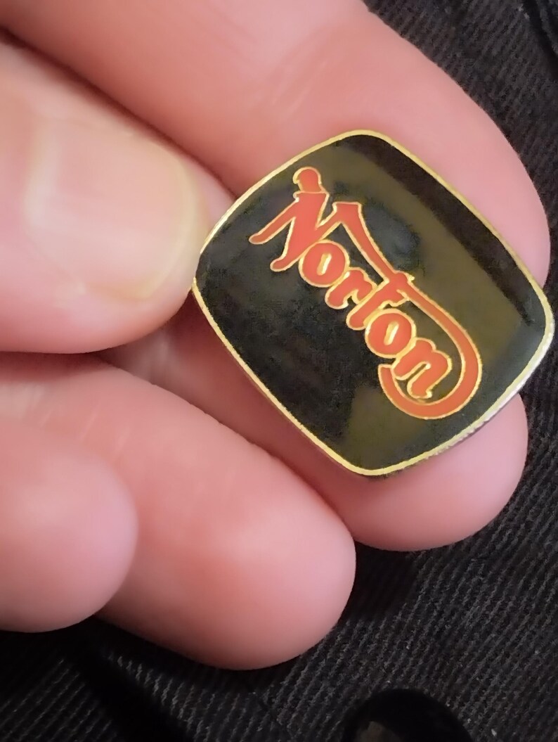 May include: A black and gold square pin with the word "Norton" in red lettering.