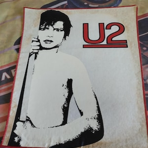 Può includere: Un'immagine in bianco e nero di una persona con i capelli corti, che tiene un oggetto lungo e sottile. Lo sfondo è bianco con la parola "U2" in lettere rosse.