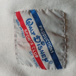 May include: A white label with the text "Walt Disney Productions Sports Club by Tiesse" in blue, and red stripes. The label is stitched onto a white fabric.