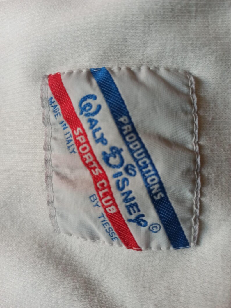 May include: A white label with the text "Walt Disney Productions Sports Club by Tiesse" in blue, and red stripes. The label is stitched onto a white fabric.