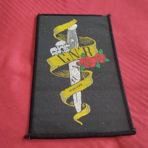May include: Black rectangular patch with a silver dagger piercing a yellow banner displaying "G N' R" in black. The design includes three skulls, red roses, and the word "DANGER" on the banner.