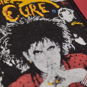 Puede incluir: Un parche rectangular de tela negra con un estampado gráfico de The Cure. El diseño incluye una figura central con cabello oscuro, una prenda roja y texto amarillo en la parte superior. Los bordes del parche tienen un acabado con costura negra.