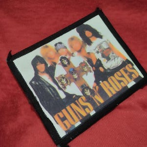 May include: A square patch with a black border, displaying a group of people. The patch features the words "GUNS N' ROSES" in orange. The image is set against a red background.