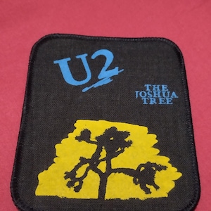 May include: Black square patch with the band name "U2" in blue, and the text "THE JOSHUA TREE" in blue. A yellow Joshua tree graphic is in the center. The patch is designed to be sewn onto clothing or bags.