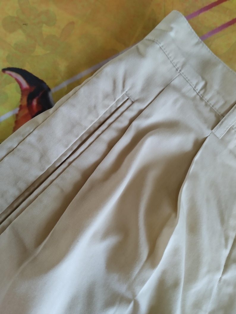Puede incluir: Primer plano de un pantal&oacute;n beige, que muestra la cintura, los pliegues y los detalles del bolsillo. La tela parece ser un material ligero, con costuras visibles a lo largo de las costuras. El pantal&oacute;n est&aacute; cuidadosamente doblado, lo que sugiere un ajuste a medida.