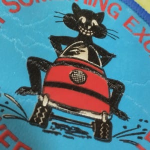 Op de afbeelding: Een patch met een zwarte cartoonkat die een rood voertuig bestuurt op een blauwe achtergrond. De kat heeft een brede grijns en witte ogen. De patch heeft de rode tekst "SOMETHING EXCITING" en "SEEN YOUR".