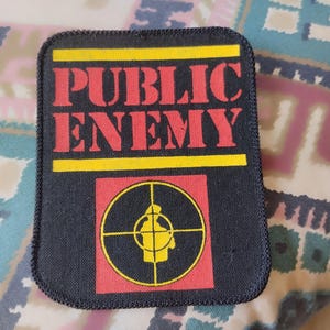 Op de afbeelding: Een rechthoekige zwarte stoffen patch met de tekst PUBLIC ENEMY in dikke rode hoofdletters tussen twee horizontale gele balken. Onder de tekst bevindt zich een rood vierkant met een geel vizier-symbool met daarin een silhouetfiguur.