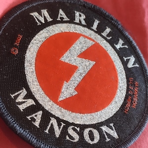 May include: Circular black patch with the name "MARILYN MANSON" in silver lettering. A red circle in the center features a silver lightning bolt. The patch has a silver border and a blue stitched edge. Copyright information is visible.