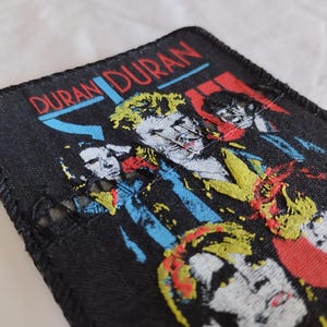 Op de afbeelding: Een rechthoekige zwarte stoffen patch met een borduursel van de band Duran Duran. Het ontwerp bevat de tekst DURAN DURAN in rood en blauw, samen met gestileerde portretten van de bandleden in geel, rood en wit.