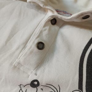 May include: Close-up of a white collared shirt with a button-up placket. The shirt features a cartoon graphic at the bottom and a black stripe on the side. The buttons are dark grey.
