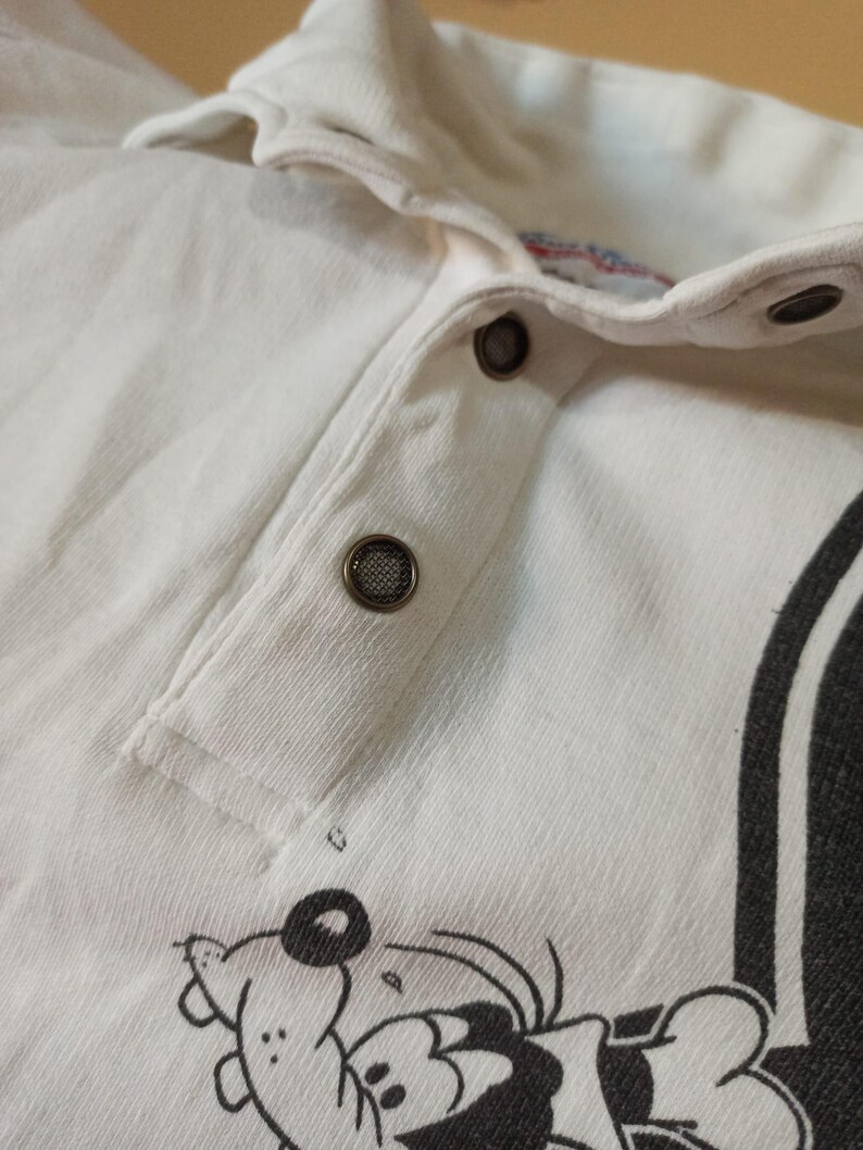 May include: Close-up of a white collared shirt with a button-up placket. The shirt features a cartoon graphic at the bottom and a black stripe on the side. The buttons are dark grey.