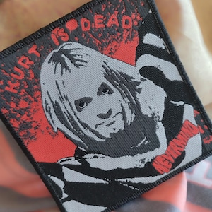 Peut inclure: Patch brodé noir et blanc avec un portrait de Kurt Cobain. Le patch a du texte rouge qui dit "Kurt is Dead" et "Nirvana" dans le coin inférieur droit.