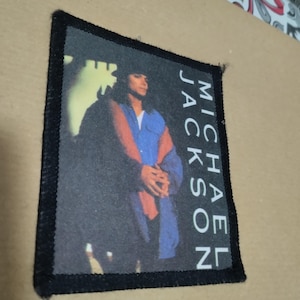 Könnte beinhalten: Ein rechteckiger Stoffaufnäher mit schwarzem gesticktem Rand, der ein gedrucktes Bild von Michael Jackson in einem blauen Hemd und einem dunklen Mantel zeigt. Der Text MICHAEL JACKSON ist vertikal in weißen Großbuchstaben auf der rechten Seite gedruckt.