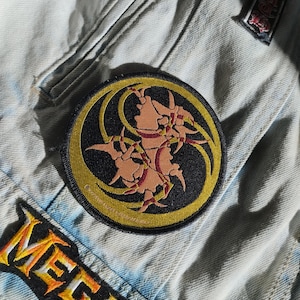 May include: Close-up of a light blue denim jacket with two patches. A round patch with a black background and a yellow border features a stylized design in shades of tan and red. Below, a patch with the word "MEGADETH" in orange and yellow.
