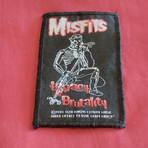 May include: Black rectangular patch with the word "Misfits" in white and red lettering. A skeleton figure with a guitar is in the center. The words "Legacy Brutality" are below the figure in red.