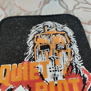Peut inclure: Un écusson rectangulaire en tissu noir présentant une illustration graphique d'un personnage masqué en orange et blanc. Le texte "QUIET RIOT" est imprimé en lettres orange grasses sur la partie inférieure du motif avec une bordure noire cousue.