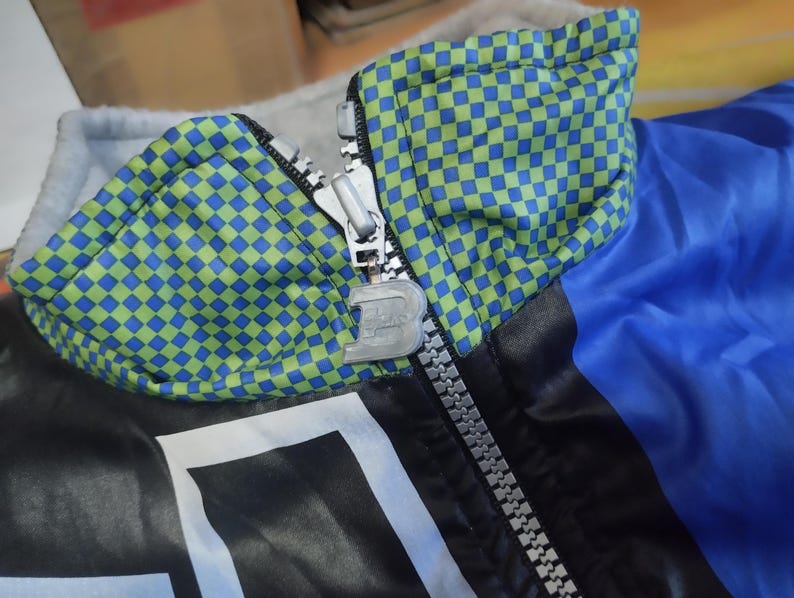 May include: A blue and black jacket with a green and blue checkered pattern on the collar. The jacket has a zipper closure and a silver zipper pull.