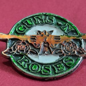 Pode incluir: Um alfinete de metal com o logotipo Guns N' Roses. O alfinete tem uma borda verde com o nome da banda em letras verdes. O centro tem um fundo branco com um desenho de arma laranja e rosas pretas.