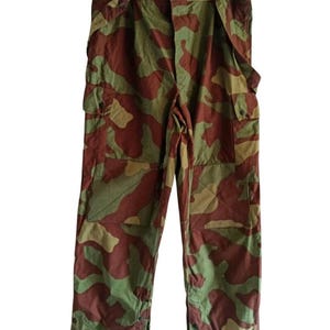 May include: Cargo trousers featuring a camouflage pattern in shades of olive green, brown, and tan. The trousers include two large side cargo pockets with button closures, reinforced knee panels, and adjustable button tabs at the waist and leg cuffs.