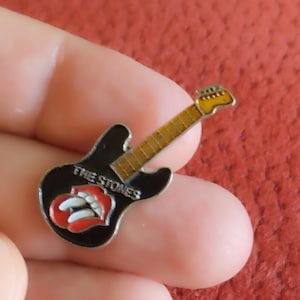 May include: A black guitar-shaped enamel pin featuring the Rolling Stones' tongue and lips logo. The pin has a gold-coloured neck and headstock, with the words "THE STONES" in white.