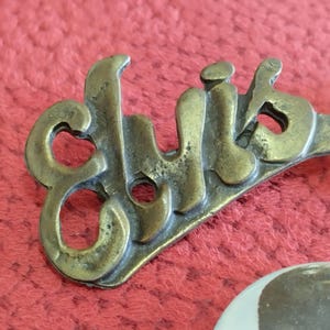 May include: A brass pin with the word "Club" written in a stylized font.