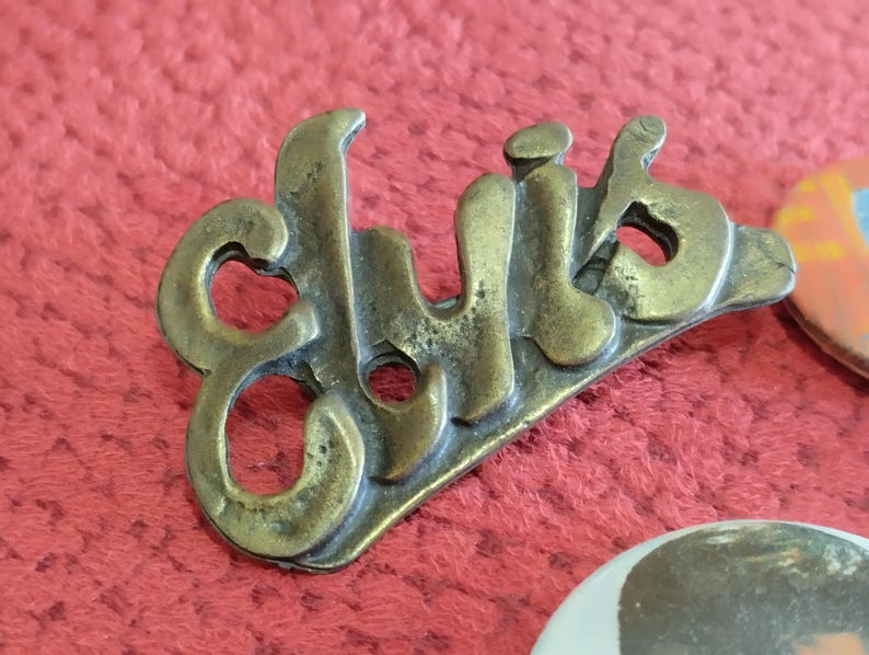 May include: A brass pin with the word "Club" written in a stylized font.