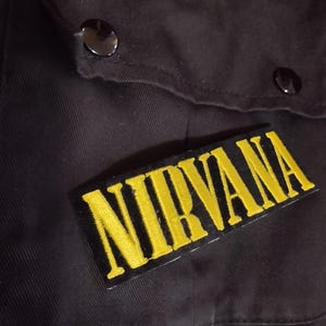 Op de afbeelding: Zwarte jas met een gele "NIRVANA" patch die op de voorkant is genaaid.