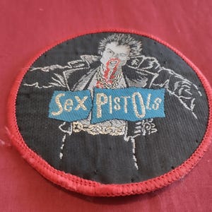 May include: Round black patch with a red stitched border featuring a graphic of a person with blood and the words "Sex Pistols" on a blue banner. The patch is likely for a jacket or bag.