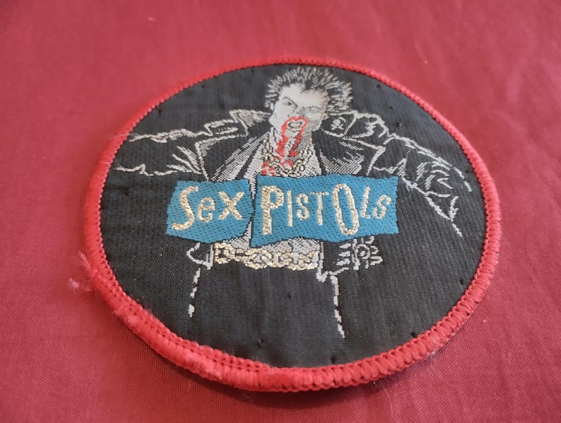 May include: Round black patch with a red stitched border featuring a graphic of a person with blood and the words "Sex Pistols" on a blue banner. The patch is likely for a jacket or bag.