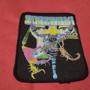Könnte beinhalten: Schwarzer Aufnäher mit dem Bandnamen "SCORPIONS" in einer farbenfrohen, stilisierten Schrift. Das Design umfasst eine Gitarre, einen Skorpion und Schädelmotive. Der Aufnäher hat einen schwarzen Rand.