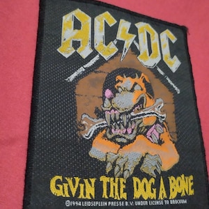 May include: Black patch featuring the AC/DC logo in yellow and white, with a cartoon dog holding a bone. The text "GIVIN THE DOG A BONE" is below the image. The patch is rectangular and has a black border.