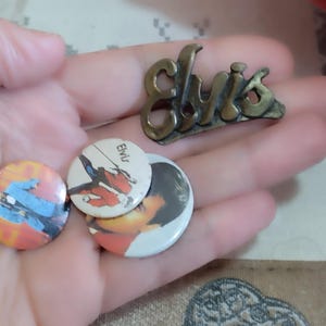 May include: Three Elvis Presley buttons, two with images of Elvis and one with the word "Elvis" in white letters on a black background. A gold metal pin with the word "Elvis" in raised letters.