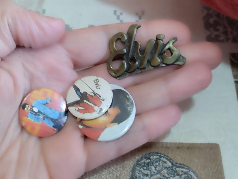 May include: Three Elvis Presley buttons, two with images of Elvis and one with the word "Elvis" in white letters on a black background. A gold metal pin with the word "Elvis" in raised letters.
