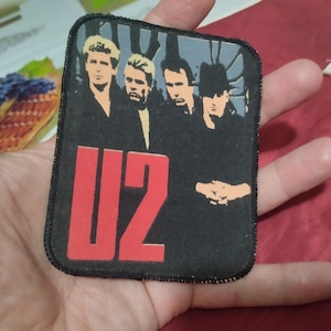 May include: A rectangular fabric patch featuring a graphic print of the band U2. The design includes the four band members in a stylized black and white illustration with the text U2 in large, bold red letters at the bottom left corner.