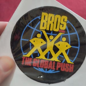 May include: A circular sticker featuring the word "BROS" in yellow above a graphic of three yellow figures. Below, in red, is "THE GLOBAL PUSH". A blue globe design forms the background of the sticker.