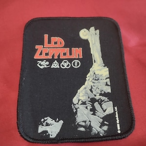 Może przedstawiać: Prostokątna czarna naszywka z czerwonym napisem Led Zeppelin, czterema białymi okrągłymi symbolami oraz białą grafiką przedstawiającą postać na szczycie skalistej góry. Naszywka posiada czarne obszycie krawędzi oraz niewielki napis.