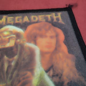 Könnte beinhalten: Ein rechteckiger Stoffaufnäher mit schwarzem gesticktem Rand, der oben das Wort MEGADETH in gelber Schrift zeigt. Die Grafik zeigt eine skelettartige Figur im Anzug neben einer Person mit langem Haar in den Farben Orange, Gelb und Schwarz.