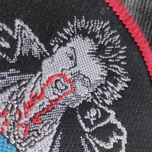 May include: Embroidered patch featuring a stylized punk rock figure with white hair and a red eye. The patch has a black background with a red border and a blue accent.