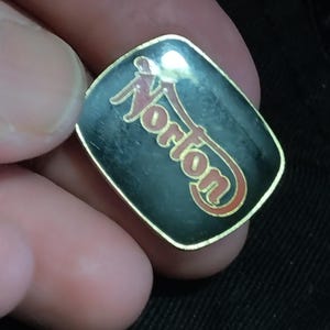 May include: A black and gold pin with the word "Norton" in red and gold lettering.