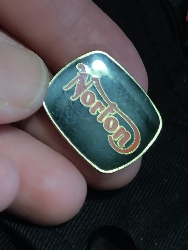 May include: A black and gold pin with the word "Norton" in red and gold lettering.