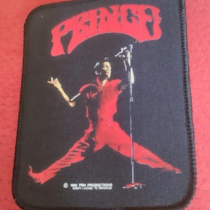 May include: Black patch with a red graphic of a performer on stage, with the name "PRINCE" arched above. The performer is in a red outfit and holding a microphone. The patch has a black border and the text "© 1999 PRIN PRODUCTIONS UNDER LICENSE TO BROCKUM".