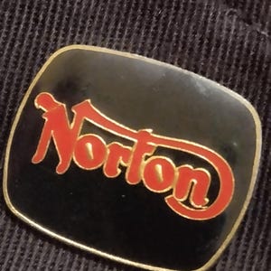 May include: A black and gold pin with the word "Norton" in red letters.