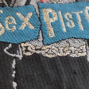 May include: Close-up of a textile with the words "Sex Pistols" in gold lettering on a turquoise blue background. The background is a dark, patterned fabric with silver and white accents. The overall aesthetic suggests a punk rock theme.